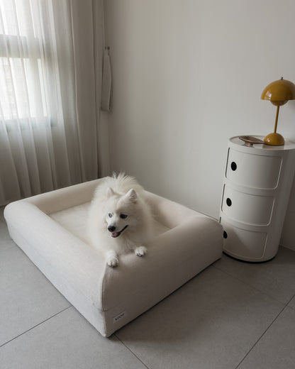A white dog resting on the bolster of the KINNE Bed by a window